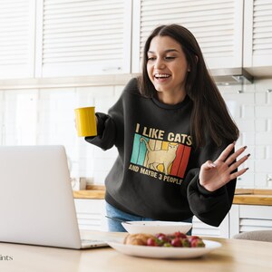 May include: A woman wearing a black sweatshirt with the text "I like cats and maybe 3 people" in a vintage style font. The sweatshirt features a retro-style graphic of a cat. She is holding a yellow mug and smiling at a laptop.