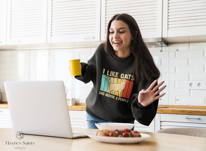 May include: A woman wearing a black sweatshirt with the text "I like cats and maybe 3 people" in a vintage style font. The sweatshirt features a retro-style graphic of a cat. She is holding a yellow mug and smiling at a laptop.