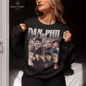 Może przedstawiać: Czarna bluza z nadrukiem przedstawiającym kilka os&oacute;b i napisem "DAN & PHIL" w stylu vintage. Widoczna jest nazwa marki "Haynes Saints Clothing Co.".