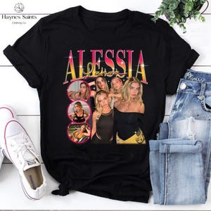 Pode incluir: T-shirt preto com o nome "Alessia" em uma fonte retrô, com um gradiente de amarelo a laranja. A camisa inclui várias imagens circulares de uma mulher. A camisa é combinada com jeans azuis e tênis brancos.