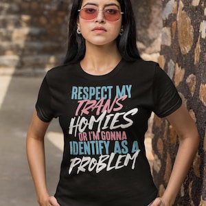 Może przedstawiać: Czarny t-shirt z napisem "RESPECT MY TRANS HOMIES OR I'M GONNA IDENTIFY AS A PROBLEM" w kolorze niebieskim, r&oacute;żowym i białym. Koszulka pochodzi od Haynes Saints Clothing Co.