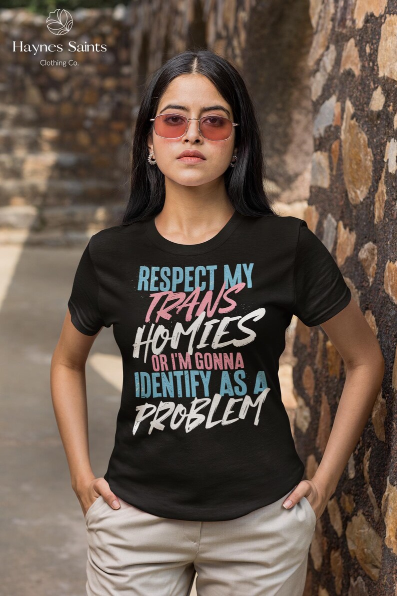 Może przedstawiać: Czarny t-shirt z napisem "RESPECT MY TRANS HOMIES OR I'M GONNA IDENTIFY AS A PROBLEM" w kolorze niebieskim, r&oacute;żowym i białym. Koszulka pochodzi od Haynes Saints Clothing Co.