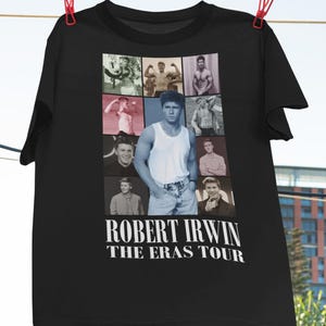 May include: Black t-shirt featuring a collage of photos of Robert Irwin at different ages. The text "Robert Irwin The Eras Tour" is printed in white below the photos.