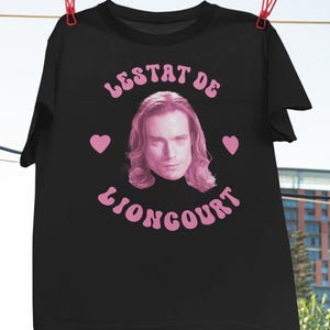 Puede incluir: Camiseta negra con un gráfico rosa de la cara de un hombre con el texto "Lestat de Lioncourt" en rosa.
