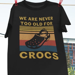 Könnte beinhalten: Schwarzes T-Shirt mit einem Vintage-Grafikdruck eines schwarzen Crocs-Schuhs. Der Text "We are never too old for Crocs" ist in Gelb auf dem Shirt gedruckt.