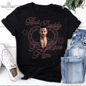 May include: Black t-shirt with the text "But Daddy I Love Him" in pink script. The design includes a photo of a man inside a heart-shaped frame with stars. The shirt is displayed with jeans and sneakers.