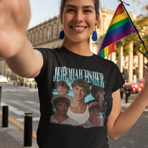 May include: Black t-shirt featuring a graphic of a group of young men with the text "Jeremiah Fisher" in a light blue font. The shirt is worn by a person holding a rainbow flag. The brand name "Haynes Saints Clothing Co." is visible.