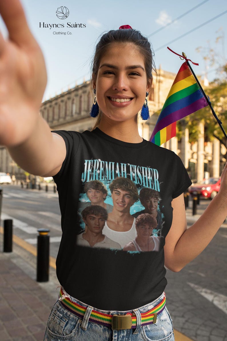 May include: Black t-shirt featuring a graphic of a group of young men with the text "Jeremiah Fisher" in a light blue font. The shirt is worn by a person holding a rainbow flag. The brand name "Haynes Saints Clothing Co." is visible.