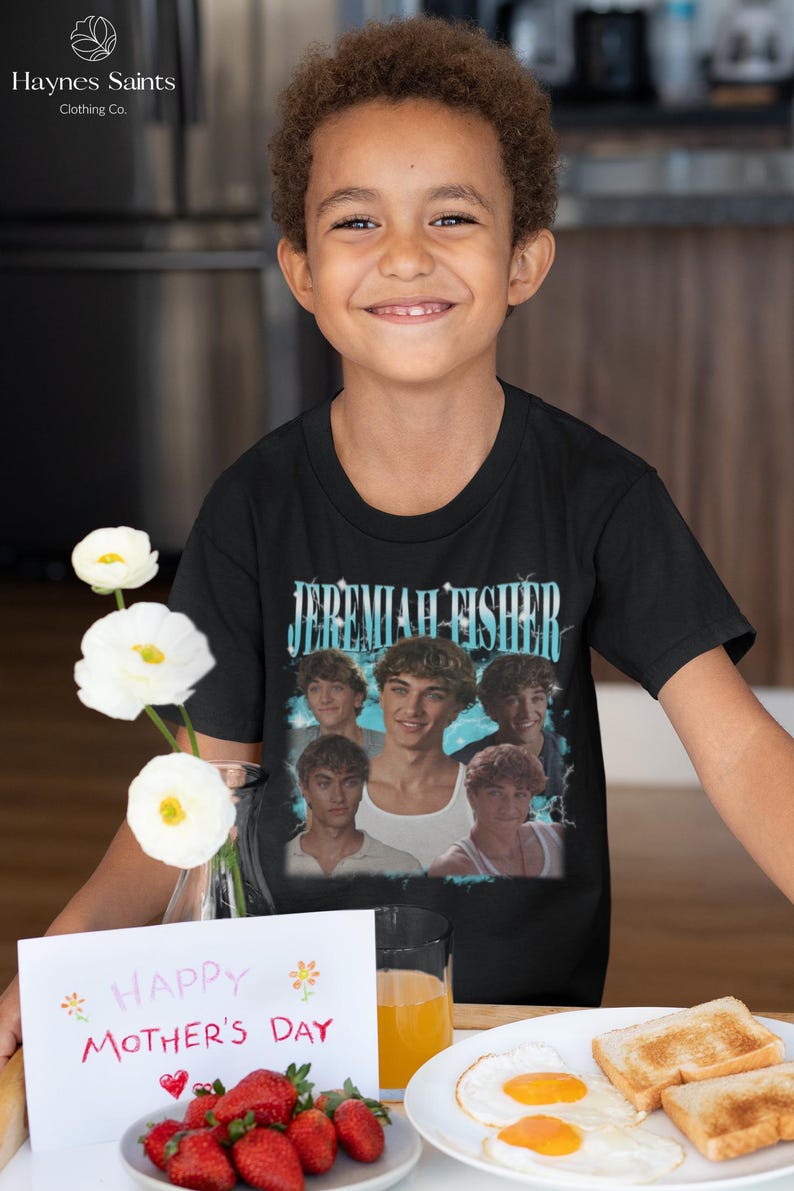 May include: A young person wearing a black t-shirt with the text "JEREMIAH FISHER" above a photo collage of young men. The image also includes a "Happy Mother's Day" card, strawberries, and a plate of eggs and toast.