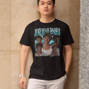 May include: Black t-shirt featuring a graphic of Jeremiah Fisher with the text "JEREMIAH FISHER" in a blue and white font. The shirt is worn by a person wearing light gray pants and a silver watch.
