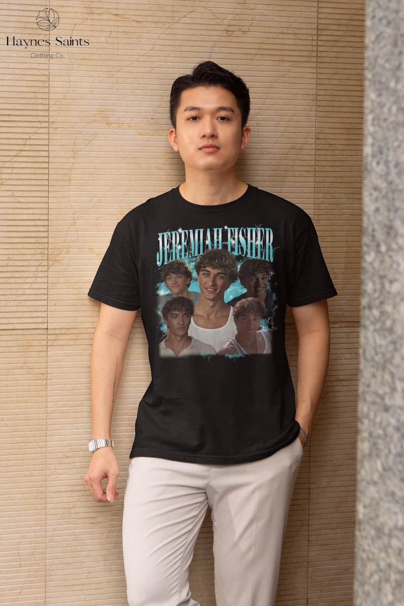 May include: Black t-shirt featuring a graphic of Jeremiah Fisher with the text "JEREMIAH FISHER" in a blue and white font. The shirt is worn by a person wearing light gray pants and a silver watch.