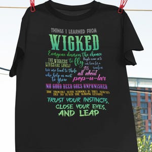 May include: Black t-shirt with the text "THINGS I LEARNED FROM WICKED" in a gradient green and blue font. The shirt features several quotes from the musical, including "TRUST YOUR INSTINCTS, CLOSE YOUR EYES, AND LEAP."