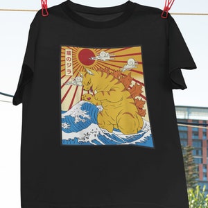 May include: Black t-shirt with a graphic print of a yellow cat monster standing on a blue wave with a red sun and rays behind it. The text "猫のジラ" is printed on the shirt.