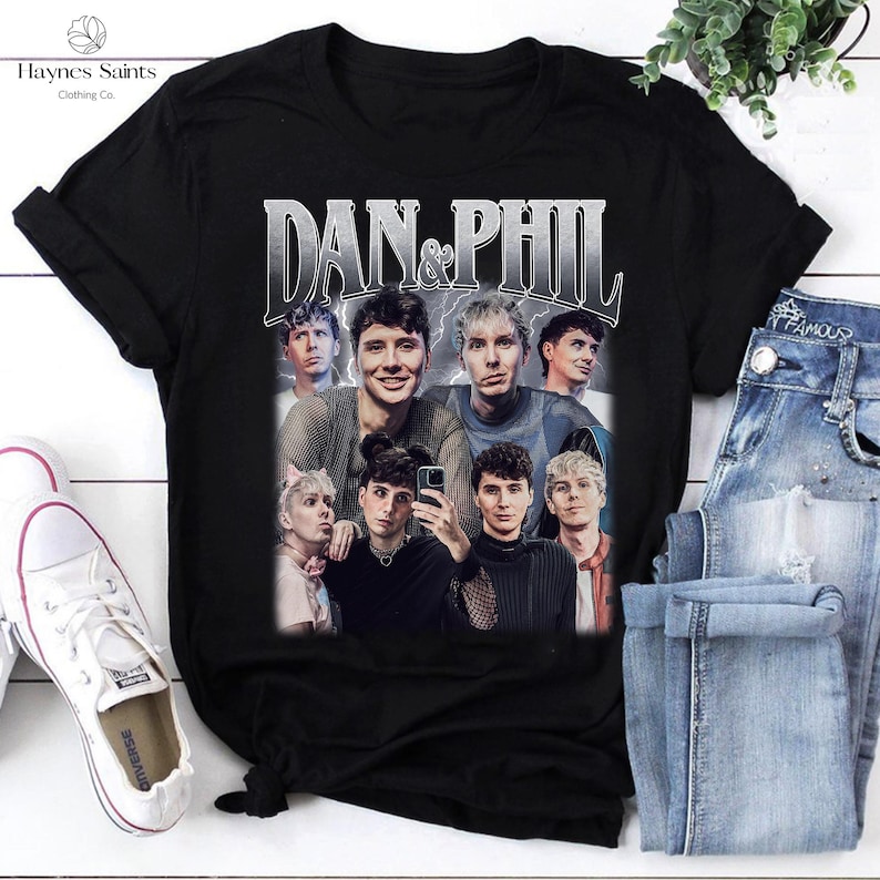 Może przedstawiać: Czarny t-shirt z grafiką w stylu vintage przedstawiającą kilka os&oacute;b i napisem "DAN & PHIL". Obraz jest ustawiony na tle niebieskich dżins&oacute;w i białych trampek. Koszulka pochodzi z Haynes Saints Clothing Co.