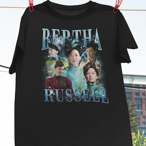 May include: Black t-shirt with a graphic of several women and the text "BERTHA RUSSELL" in a blue, vintage-style font. The image is a portrait of the women in period clothing. The shirt is hanging from a clothesline with red pegs.
