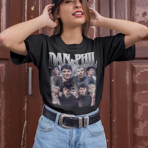 Może przedstawiać: Czarny t-shirt z grafiką przedstawiającą wiele os&oacute;b i napisem "DAN & PHIL" w srebrnym, vintage stylu. Koszulka jest noszona z niebieskimi szortami dżinsowymi i czarnym paskiem ze srebrną klamrą. Widoczna jest nazwa marki "Haynes Saints Clothing Co.".