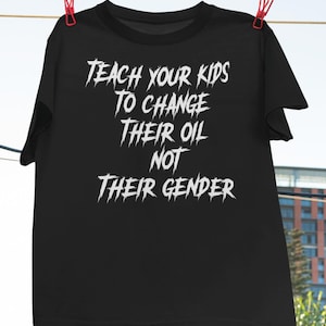 Könnte beinhalten: Schwarzes T-Shirt mit dem weißen Text "TEACH YOUR KIDS TO CHANGE THEIR OIL NOT THEIR GENDER", das an roten Wäscheklammern hängt. Das Logo von Haynes Saints Clothing Co. befindet sich in der oberen linken Ecke.