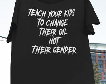 Op de afbeelding: Zwart T-shirt met de tekst "TEACH YOUR KIDS TO CHANGE THEIR OIL NOT THEIR GENDER" in het wit, hangend aan rode wasknijpers. Het logo van Haynes Saints Clothing Co. staat in de linkerbovenhoek.