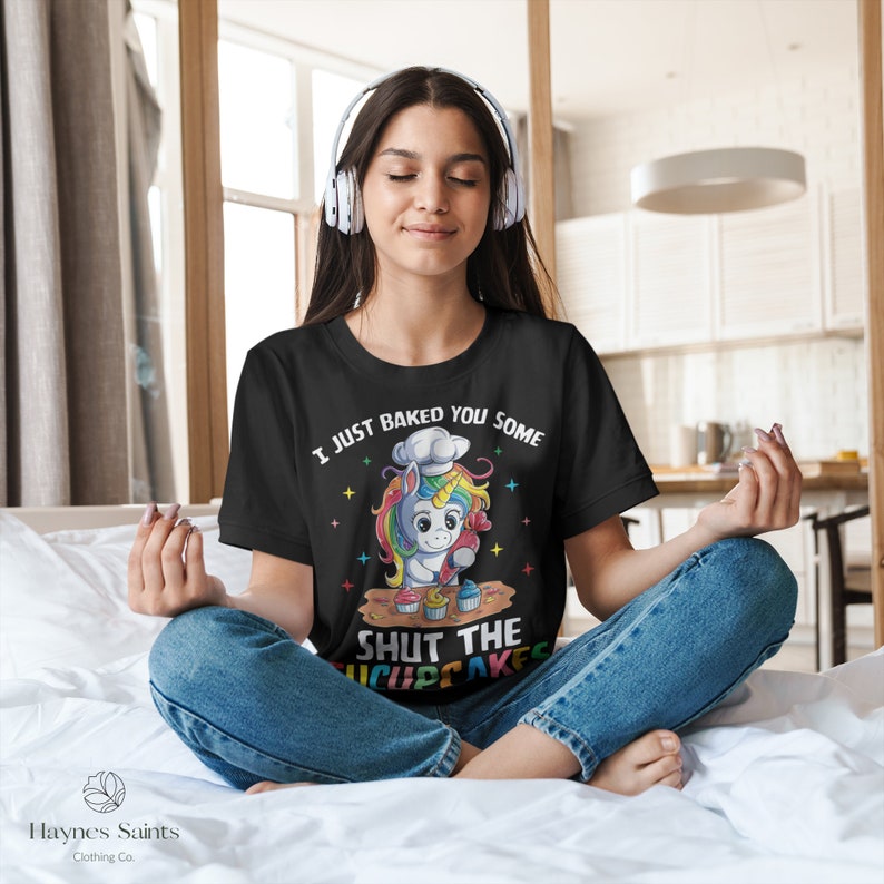 May include: A black t-shirt featuring a colourful unicorn wearing a chef's hat and holding a cupcake. The text on the shirt reads "I just baked you some shut the cupcakes".