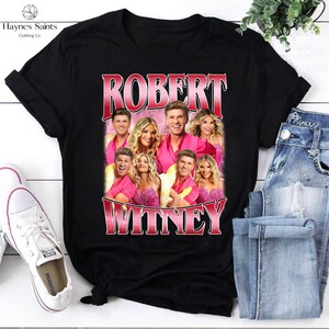 May include: Black t-shirt featuring a group photo with the names "ROBERT" and "WITNEY" in a red and pink gradient font. The shirt is from "Haynes Saints Clothing Co."