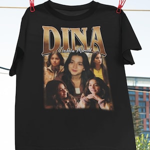 May include: Black t-shirt featuring a graphic print of "DINA" in gold lettering with "Isabela Merced" underneath. The graphic also includes four images of Isabela Merced.