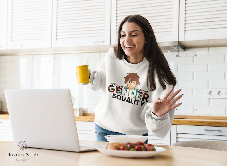 May include: A white sweatshirt featuring a colourful graphic design that reads "Advocate of True Gender Equality". The graphic also includes a cartoon illustration of a person with brown hair. The sweatshirt is being worn by a person in a kitchen setting.