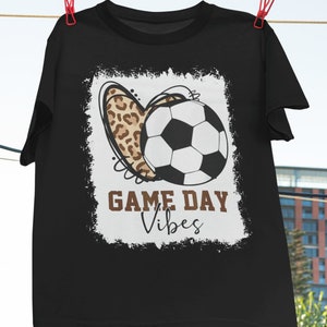 May include: Black t-shirt with a white graphic of a soccer ball and a leopard print heart. The graphic has a distressed look and the text "GAME DAY Vibes" is written below the image.