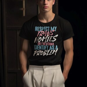 Może przedstawiać: Czarny t-shirt z napisem "RESPECT MY TRANS HOMIES OR I'M GONNA IDENTIFY AS A PROBLEM" w kolorze niebieskim, r&oacute;żowym i białym. Koszulka jest połączona z jasnymi spodniami. Widoczna jest marka "Haynes Saints Clothing Co.".