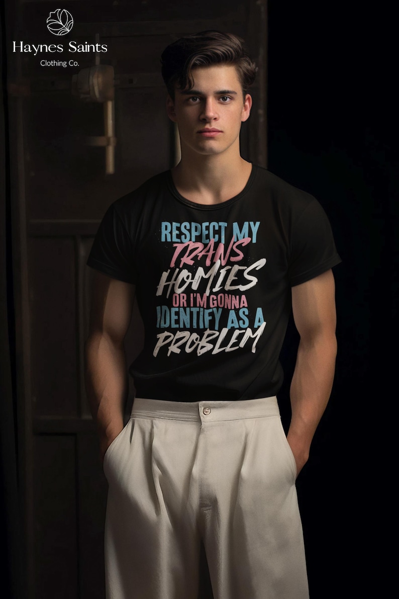 Może przedstawiać: Czarny t-shirt z napisem "RESPECT MY TRANS HOMIES OR I'M GONNA IDENTIFY AS A PROBLEM" w kolorze niebieskim, r&oacute;żowym i białym. Koszulka jest połączona z jasnymi spodniami. Widoczna jest marka "Haynes Saints Clothing Co.".