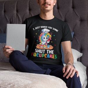 May include: A black t-shirt featuring a colourful unicorn graphic baking cupcakes. The text on the shirt reads "I just baked you some shut the fucupcakes".