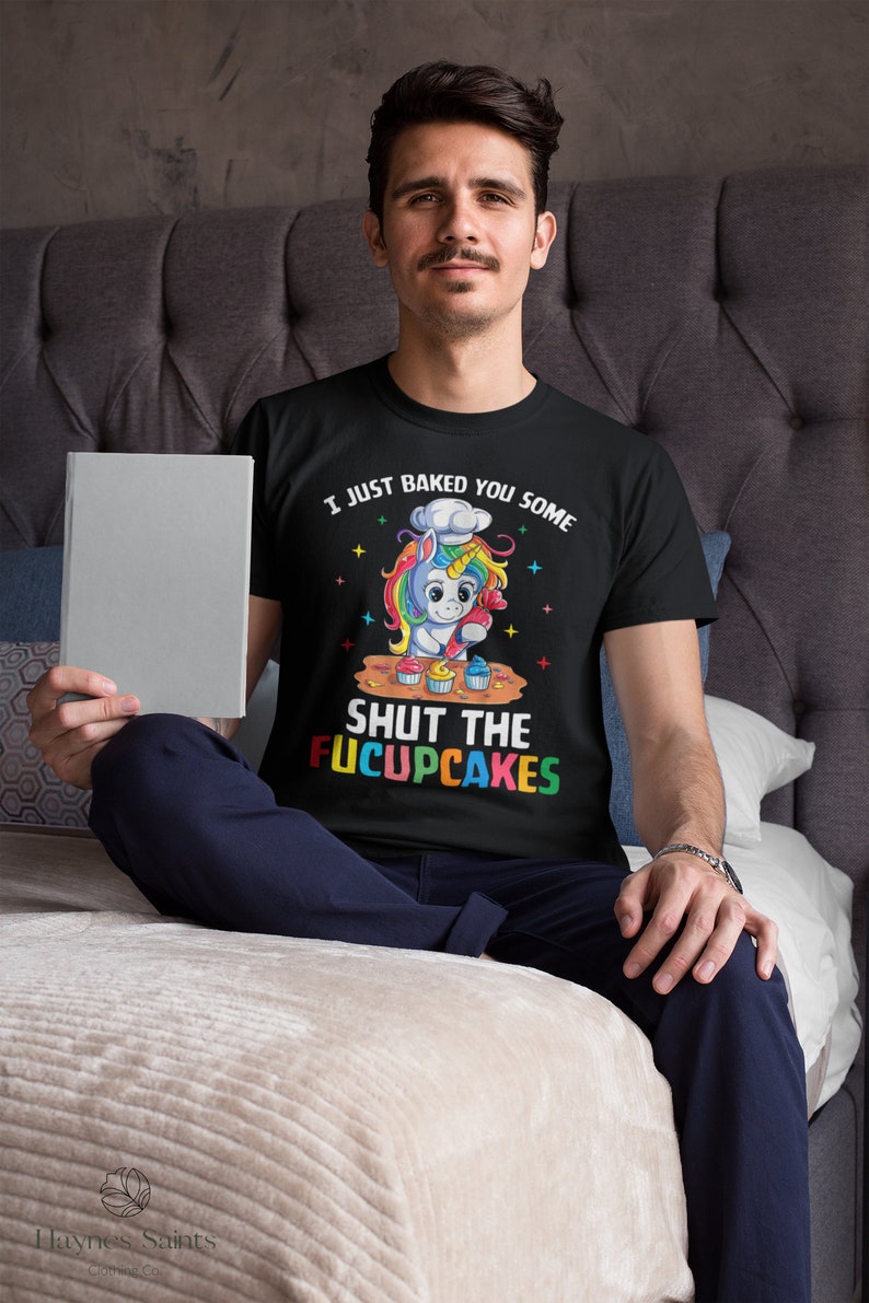 May include: A black t-shirt featuring a colourful unicorn graphic baking cupcakes. The text on the shirt reads "I just baked you some shut the fucupcakes".