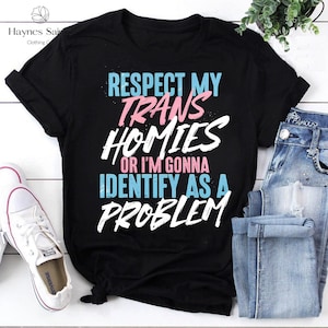Może przedstawiać: Czarny t-shirt z napisem "RESPECT MY TRANS HOMIES OR I'M GONNA IDENTIFY AS A PROBLEM" w kolorze białym, r&oacute;żowym i niebieskim. Koszulka jest zestawiona z niebieskimi dżinsami i białymi trampkami.