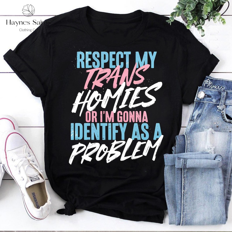 Może przedstawiać: Czarny t-shirt z napisem "RESPECT MY TRANS HOMIES OR I'M GONNA IDENTIFY AS A PROBLEM" w kolorze białym, r&oacute;żowym i niebieskim. Koszulka jest zestawiona z niebieskimi dżinsami i białymi trampkami.