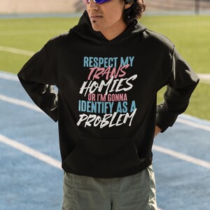 Może przedstawiać: Czarna bluza z kapturem z napisem "RESPECT MY TRANS HOMIES OR I'M GONNA IDENTIFY AS A PROBLEM" w kolorze niebieskim, r&oacute;żowym i białym. Bluza z kapturem jest noszona przez osobę w okularach przeciwsłonecznych i szortach. Nazwa marki "Haynes Saints Clothing Co." znajduje się w lewym g&oacute;rnym rogu.