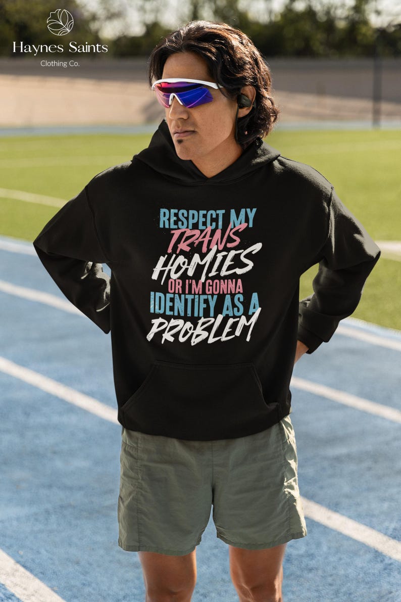 Może przedstawiać: Czarna bluza z kapturem z napisem "RESPECT MY TRANS HOMIES OR I'M GONNA IDENTIFY AS A PROBLEM" w kolorze niebieskim, r&oacute;żowym i białym. Bluza z kapturem jest noszona przez osobę w okularach przeciwsłonecznych i szortach. Nazwa marki "Haynes Saints Clothing Co." znajduje się w lewym g&oacute;rnym rogu.