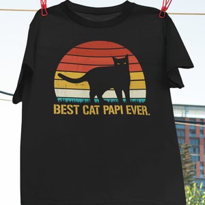 May include: Black t-shirt with a retro sunset graphic featuring a black cat silhouette. The text "BEST CAT PAPI EVER." is printed below the graphic.