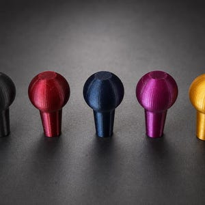 May include: Five spherical knobs with tapered bases arranged in a horizontal row. The knobs are colored in metallic grey, red, dark blue, magenta, and gold. Each piece features a smooth, rounded top and a straight, narrowing stem at the bottom.