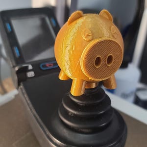 May include: A 3D-printed, golden-yellow pig figurine with a textured snout and small ears. The pig is perched atop a black joystick, showcasing a unique blend of technology and whimsical design. The figurine has a round body and four small legs.