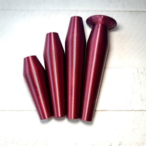 May include: Four metallic burgundy-colored bottle stoppers of varying sizes are arranged on a white wooden surface. The stoppers have a tapered design, with one featuring a flat, circular top.