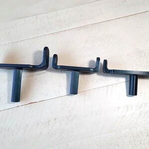 May include: Three dark blue metal wall hooks are arranged horizontally against a white wooden background. Each hook has a flat base and a curved top, with a cylindrical post connecting the base to the wall. The hooks are of varying sizes.