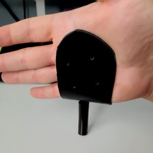 May include: A black, curved, leather-like object with small holes, attached to a black cylindrical base. The object is held in a hand, with the base resting on a white surface.