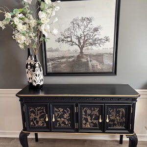May include: A black sideboard with gold accents and floral designs. The cabinet has four doors with gold handles and sits on curved legs. Above the sideboard is a framed black and white tree landscape print. Two black and white vases with white flowers are on the left.