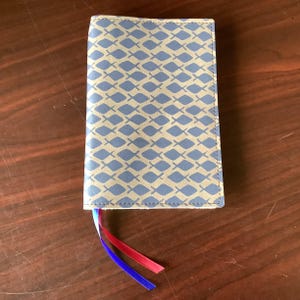 May include: A closed, rectangular book cover with a blue and tan fish pattern. The cover has three colored ribbon bookmarks: red, blue, and white. The cover is made of a leather-like material.