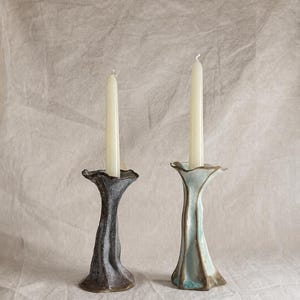 May include: Two tall, slender ceramic candle holders with organic, rippled shapes. One holder is dark charcoal grey with a textured finish, while the other is light blue with brown edges. Each holds a single, plain, off-white taper candle.