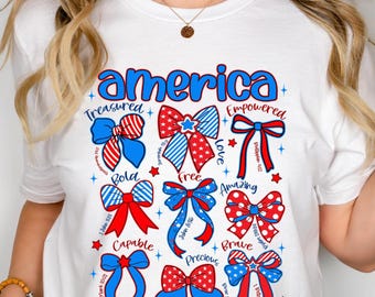 May include: White t-shirt with a patriotic design featuring the word "America" in blue, along with various bow illustrations in red, white, and blue. The bows are accompanied by words like "Treasured," "Bold," and "Capable."