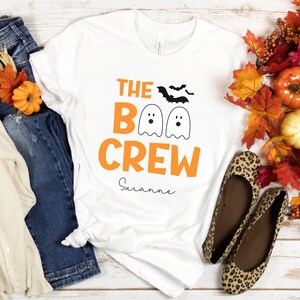 May include: White t-shirt with orange text that reads "The Boo Crew" and a personalized name "Suzanne". The shirt features three cartoon ghosts with black eyes and mouths. There are also two black bats flying above the ghosts.