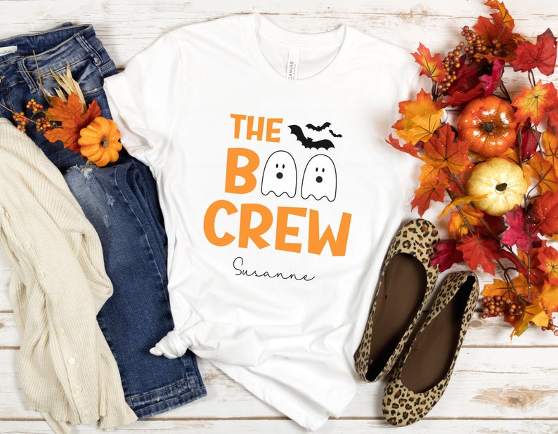 May include: White t-shirt with orange text that reads "The Boo Crew" and a personalized name "Suzanne". The shirt features three cartoon ghosts with black eyes and mouths. There are also two black bats flying above the ghosts.