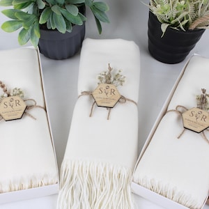 Pode incluir: Lenços cor marfim com franjas, apresentados em caixas brancas. Cada lenço é adornado com uma etiqueta de madeira com iniciais e detalhes florais, amarrada com barbante. Plantas em vasos são visíveis ao fundo.