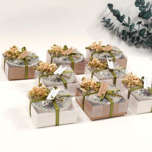 Op de afbeelding: Een arrangement van geschenkdozen, elk met een decoratieve kom, versierd met gedroogde bloemen en een lint. De kommen hebben een blauw-wit bloemenpatroon. De dozen zijn in bruin- en wittinten.