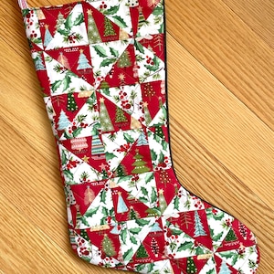 May include: A quilted Christmas stocking with a festive design. The stocking features a patchwork pattern of red, white, and green triangles, adorned with Christmas trees, holly, and berries. A red and white striped loop is attached for hanging.